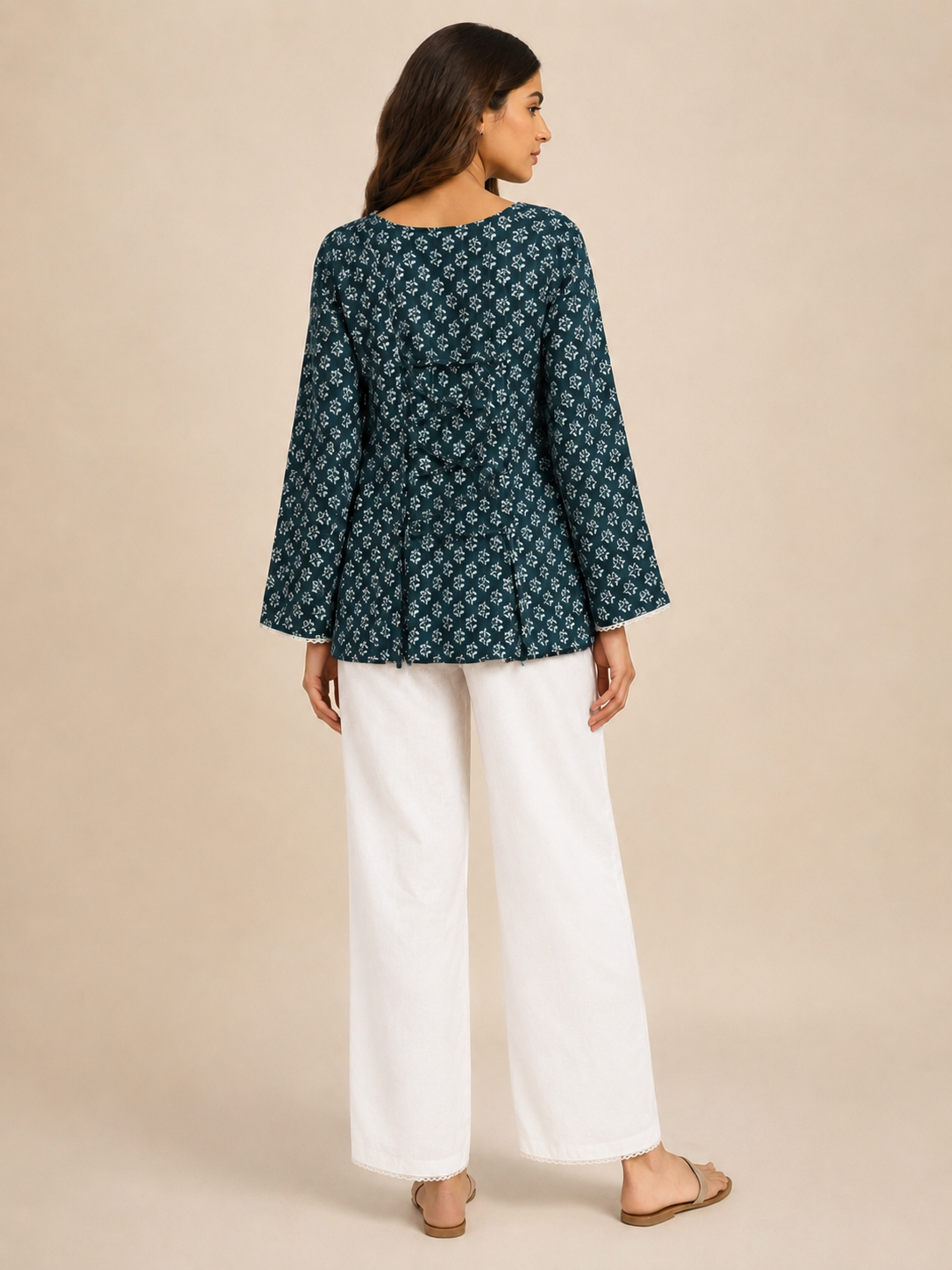 A woman is shown from the back wearing a dark teal long-sleeve kurta with a white floral pattern, paired with white wide-leg trousers and flat sandals, set against a solid beige studio background.