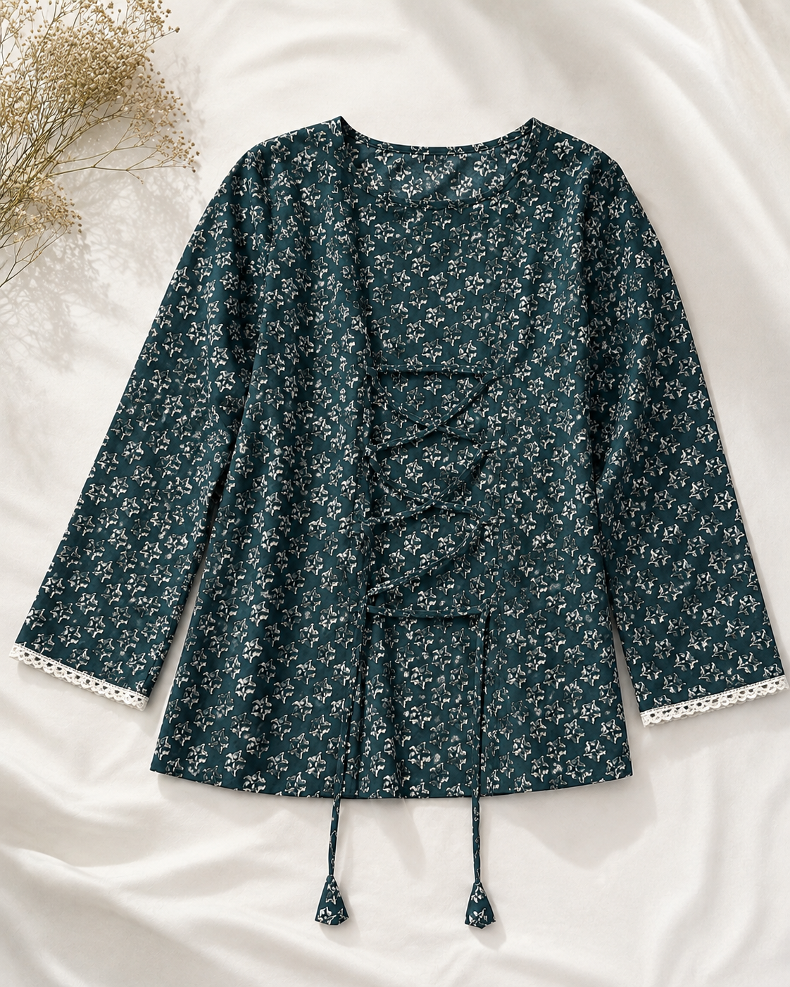 A dark teal floral long-sleeve top with lace-up front details and tassel ties, laid flat on a white fabric background with dried flowers in the corner.