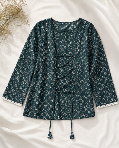 A dark teal floral long-sleeve top with lace-up front details and tassel ties, laid flat on a white fabric background with dried flowers in the corner.