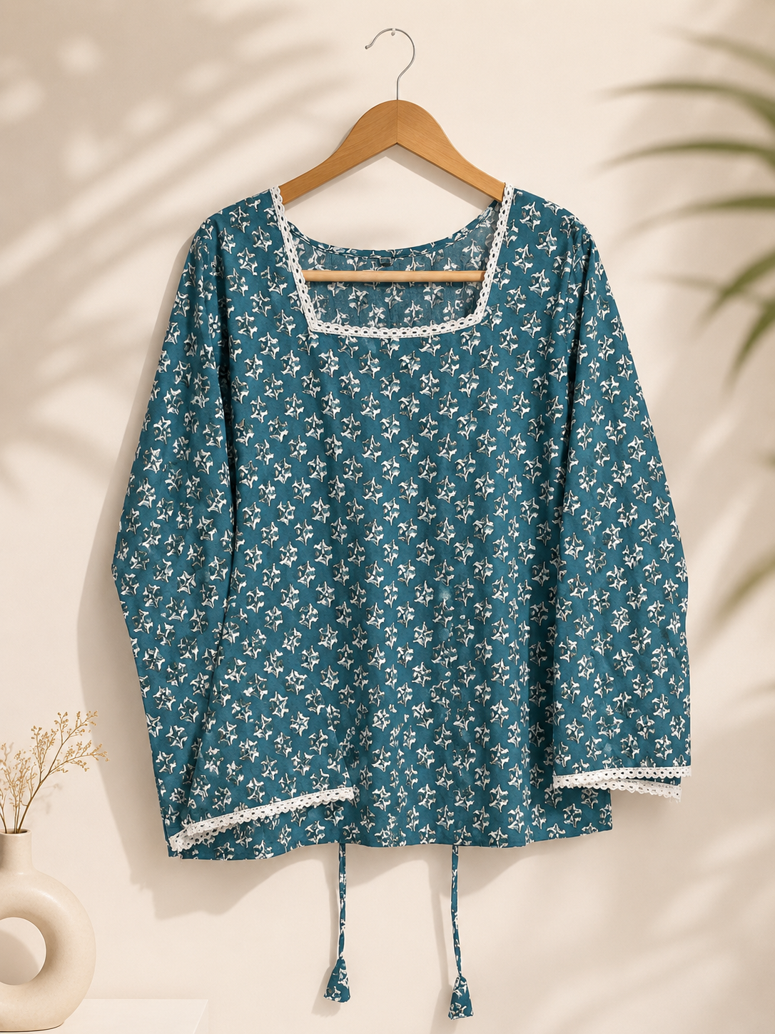 A teal blue long-sleeve top with a white floral print, hanging on a wooden hanger against a warm beige wall with soft leaf shadows.