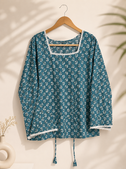 A teal blue long-sleeve top with a white floral print, hanging on a wooden hanger against a warm beige wall with soft leaf shadows.